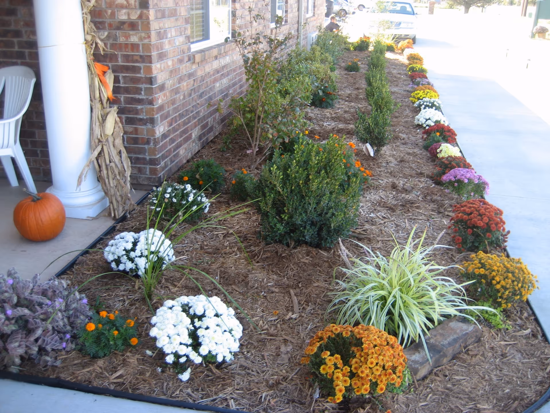 A landscaped garden bed along the side of a brick building with various colorful flowers and shrubs planted in mulch. There is a white column with dried corn stalks tied to it and a pumpkin on the concrete porch area. A white plastic chair is partially visible on the left side. A sidewalk runs alongside the garden bed, and a white vehicle is parked in the background.
