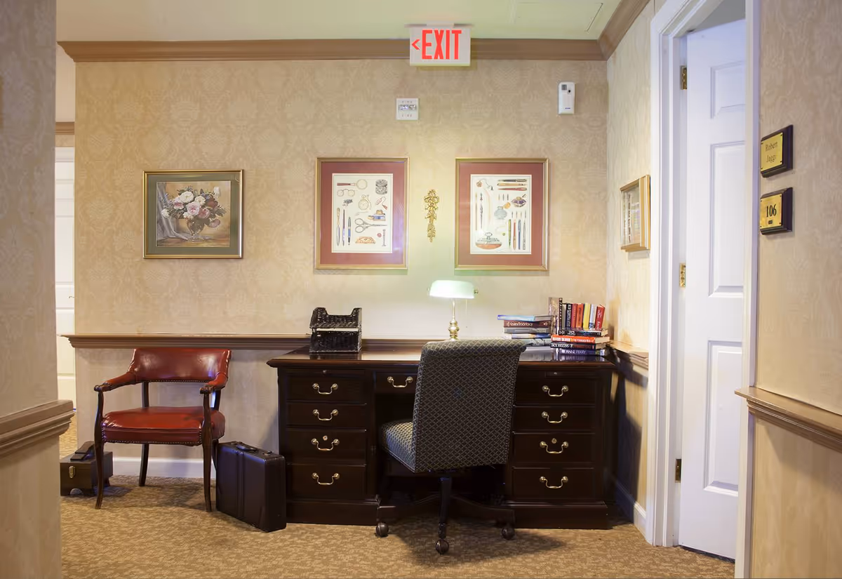 A small office or study area in a hallway with a dark wooden desk, a cushioned office chair, and a red leather armchair. The desk has a small lamp, a stack of books, and an organizer. The walls are decorated with framed artwork and wallpaper with a subtle pattern. There is an exit sign above the desk and a door labeled 106 to the right.