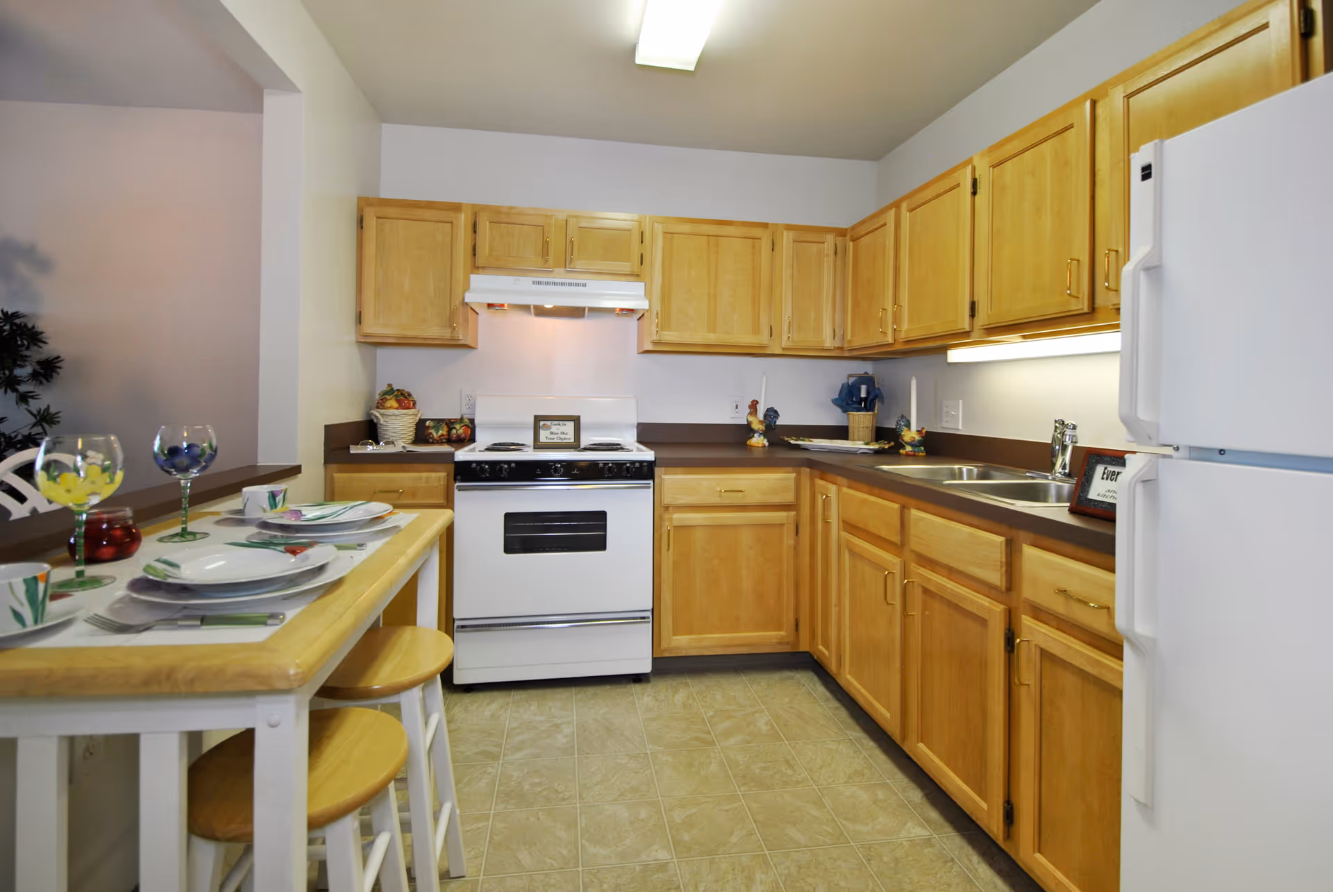 A kitchen with light wood cabinets, a white stove with a range hood, a white refrigerator, and a double sink. There is a small dining counter with two wooden stools set with plates, cups, and glasses. The floor is tiled and the walls are painted white.