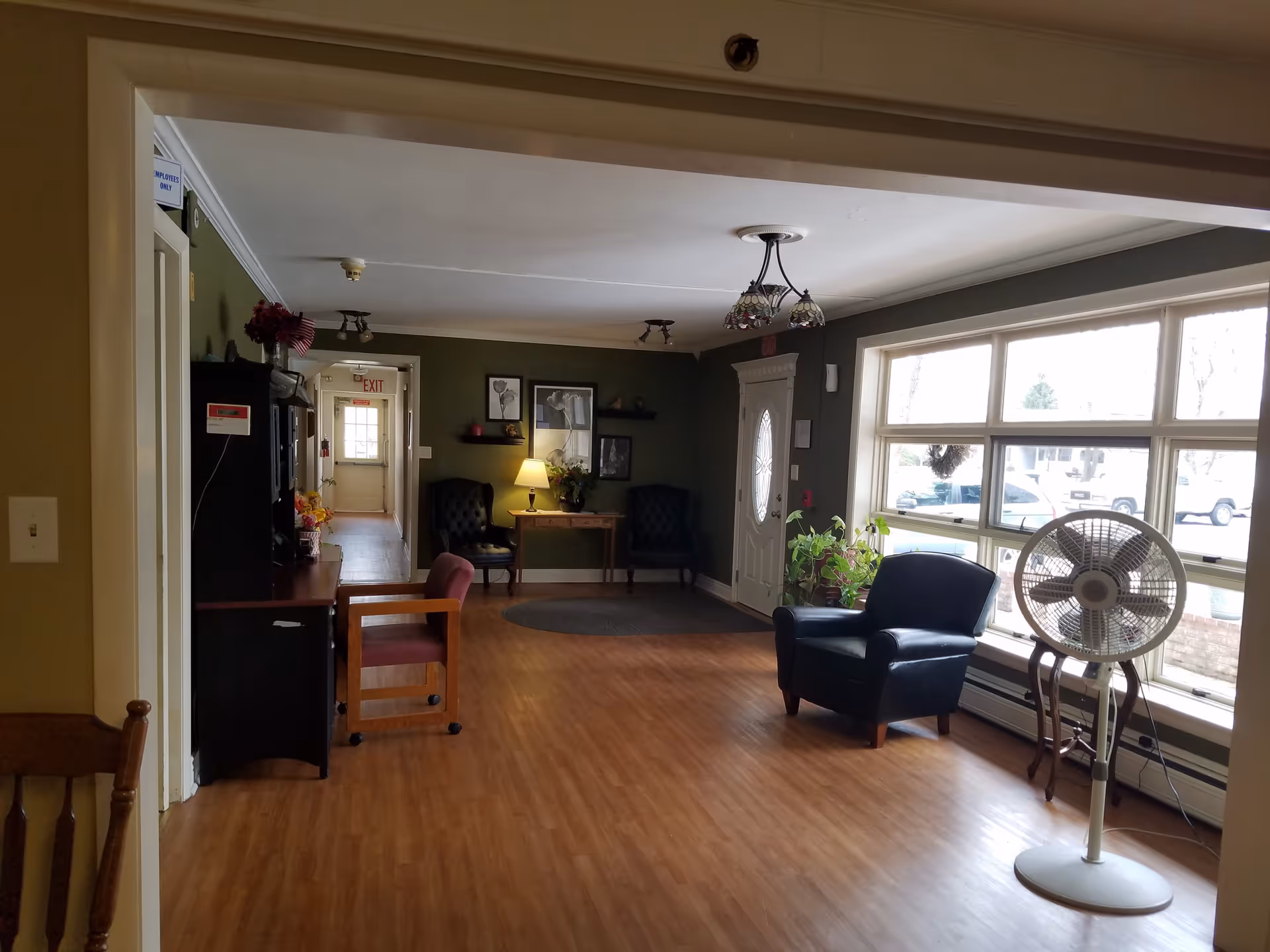 A cozy living room area with wooden flooring, two black armchairs near a small table with a lamp, a large window letting in natural light, a standing fan, and some indoor plants. The walls are painted green and decorated with framed pictures and shelves. There is a hallway leading to an exit door in the background.