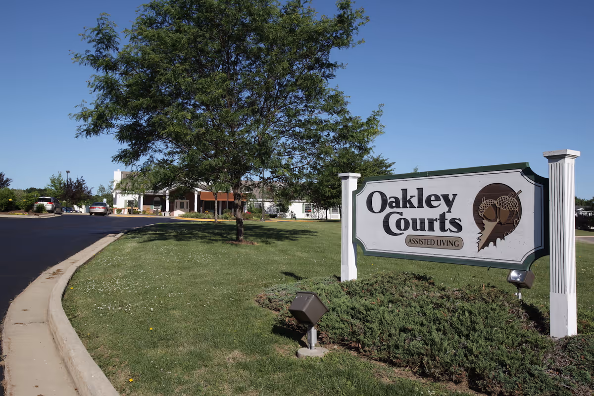 Entrance sign reading "Oakley Courts Assisted Living" on a lawn with a driveway and the facility building in the background.