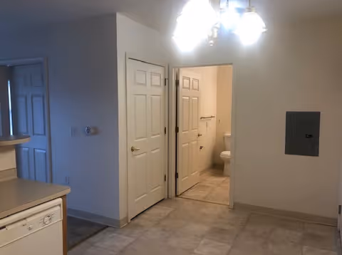 Interior view of a senior apartment showing a kitchen area with a dishwasher on the left, a hallway with two closed doors, and an open door leading to a bathroom with a visible toilet. The floor is tiled, and a ceiling light fixture is visible.