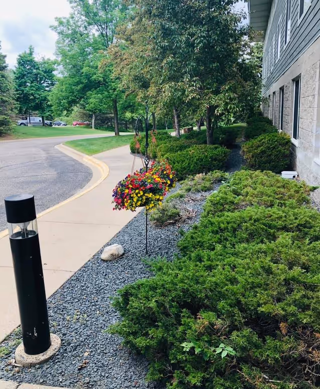 Landscaped walkway beside a senior living building with hanging flower baskets, shrubs, a bollard light, and a curved driveway.