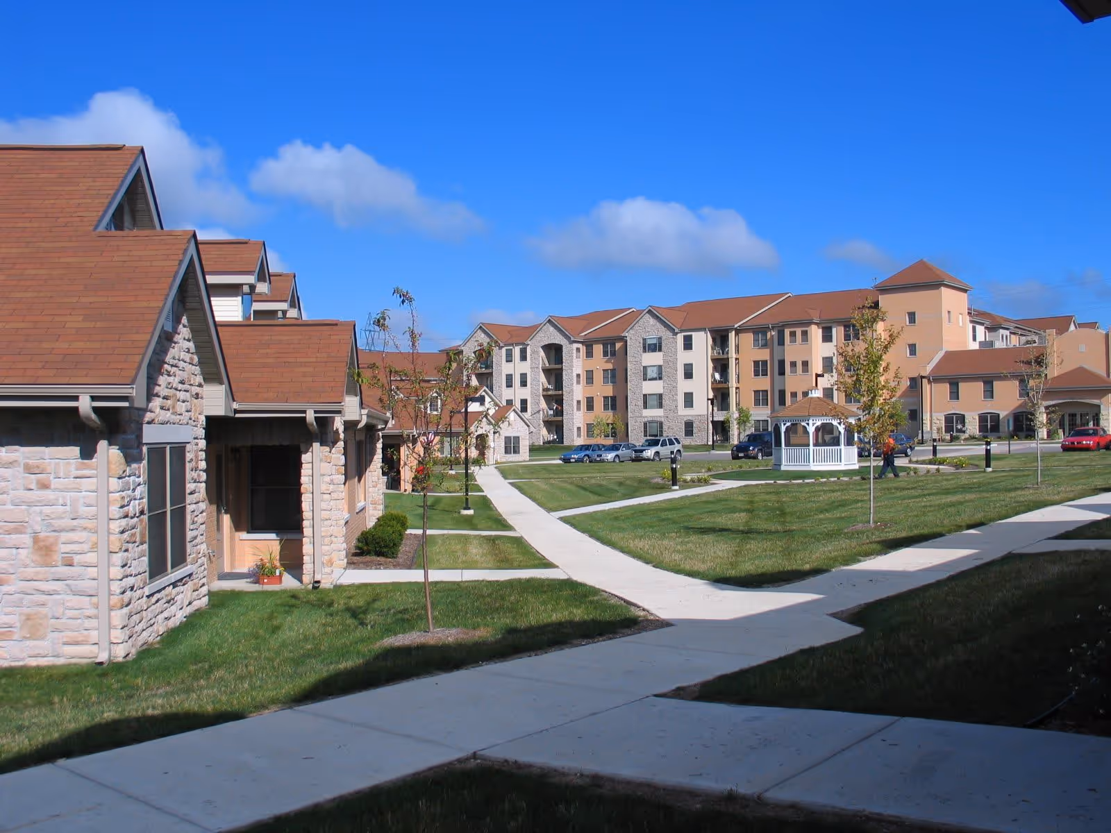 Outdoor view of Wilson Commons senior living facility showing multiple buildings with stone and beige exteriors, red roofs, a white gazebo, green lawns, young trees, and a clear blue sky.