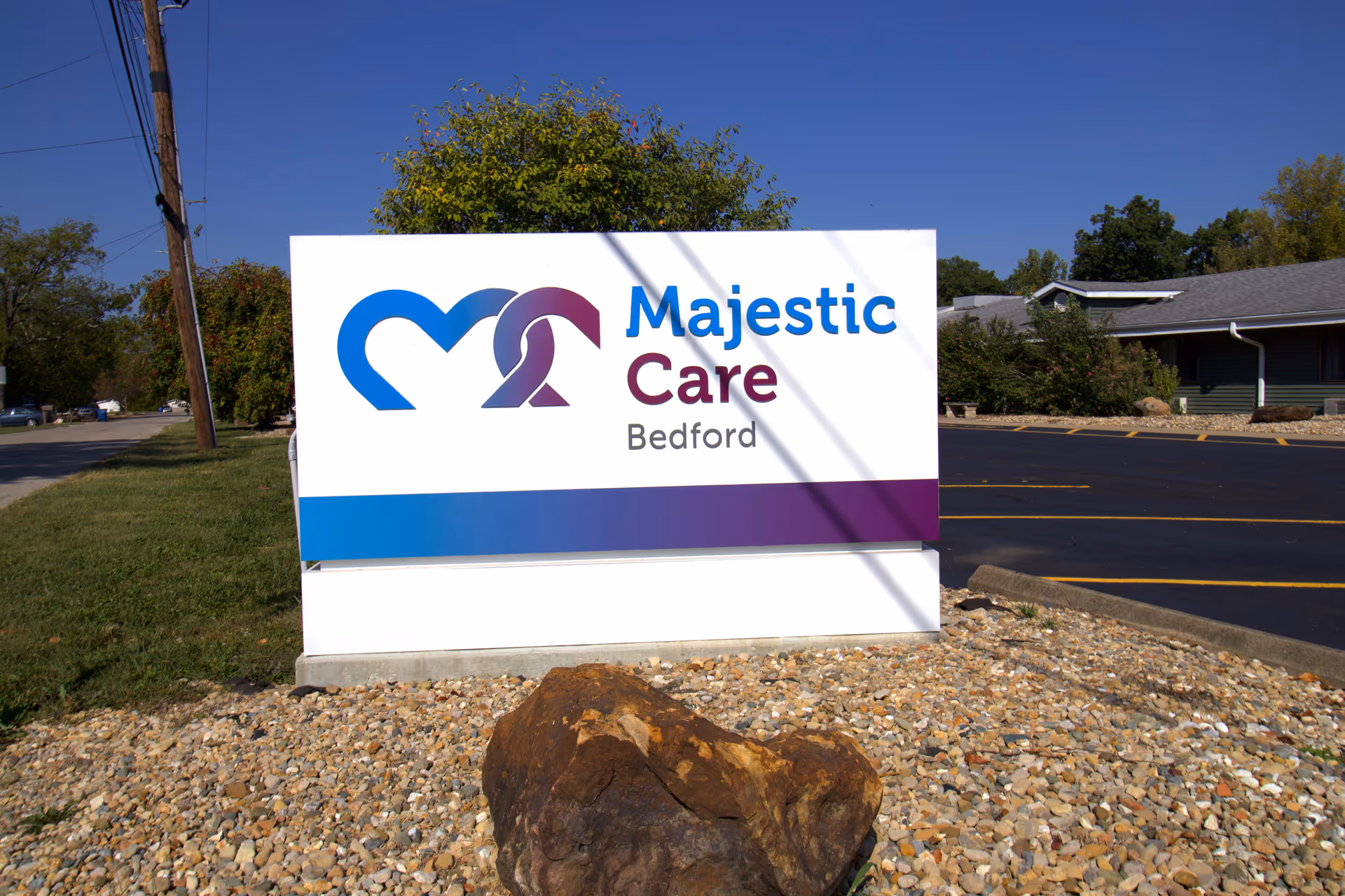 Outdoor view of a white sign with blue and purple accents that reads 'Majestic Care Bedford' placed on a bed of rocks with a large rock in front. There is a parking lot and a building in the background under a clear blue sky.