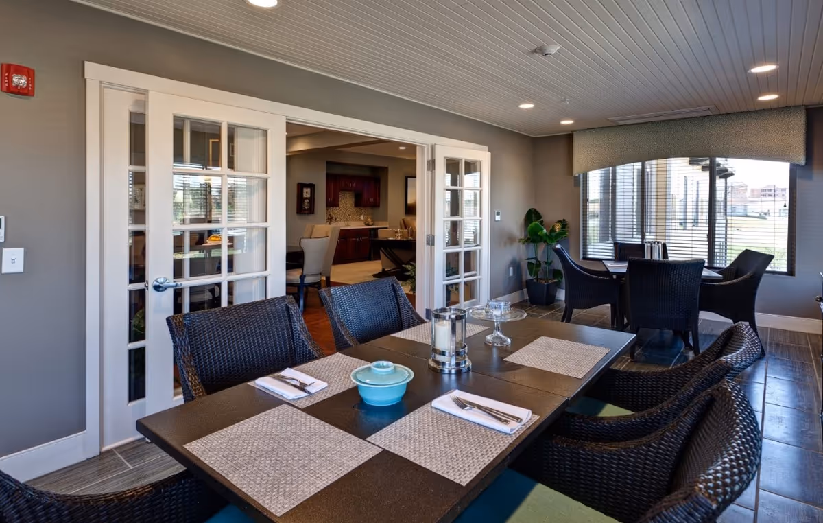 Dining room with a dark rectangular table set with placemats and wicker chairs, glass‑paneled doors, and large windows overlooking the outside.