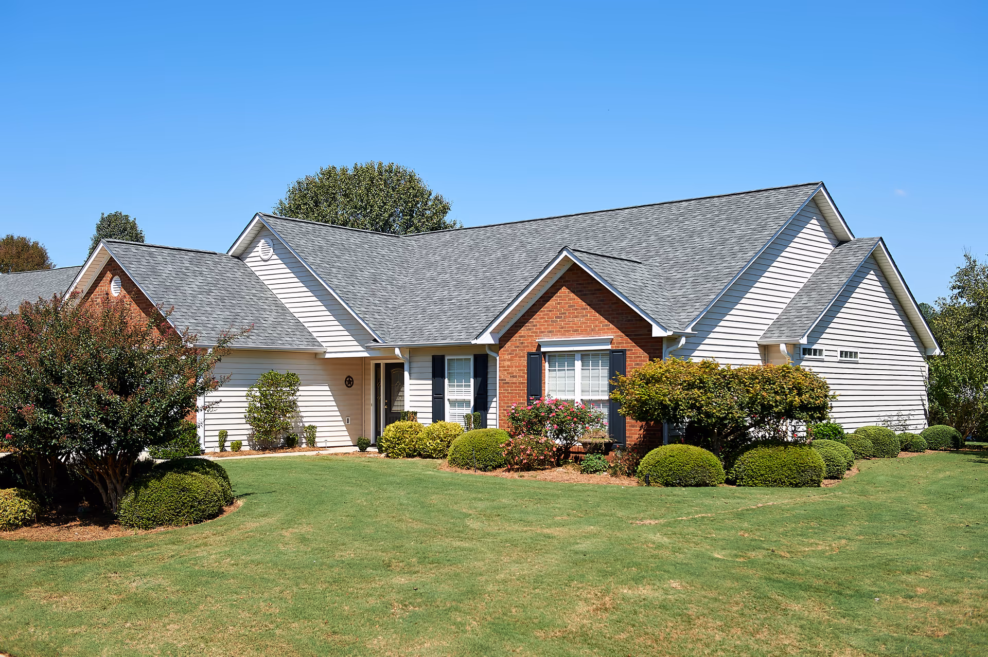 Single-story residential building with white siding and red brick accents, surrounded by neatly trimmed bushes and a well-maintained lawn under a clear blue sky.