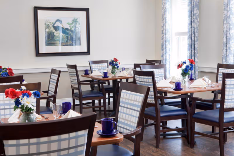 Bright dining room with multiple wooden tables and upholstered chairs set with blue cups and floral centerpieces.