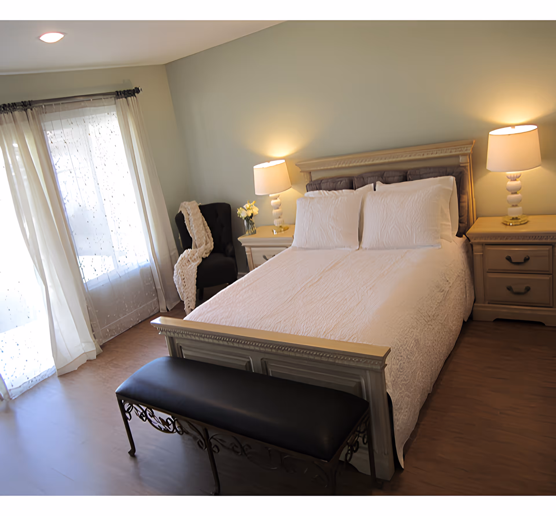 A cozy bedroom with a neatly made bed featuring white bedding and pillows. There are two bedside tables with matching lamps on each side of the bed. A black cushioned bench is placed at the foot of the bed. To the left, there is a window with sheer white curtains allowing natural light to enter the room. A black armchair with a white throw blanket is positioned near the window.