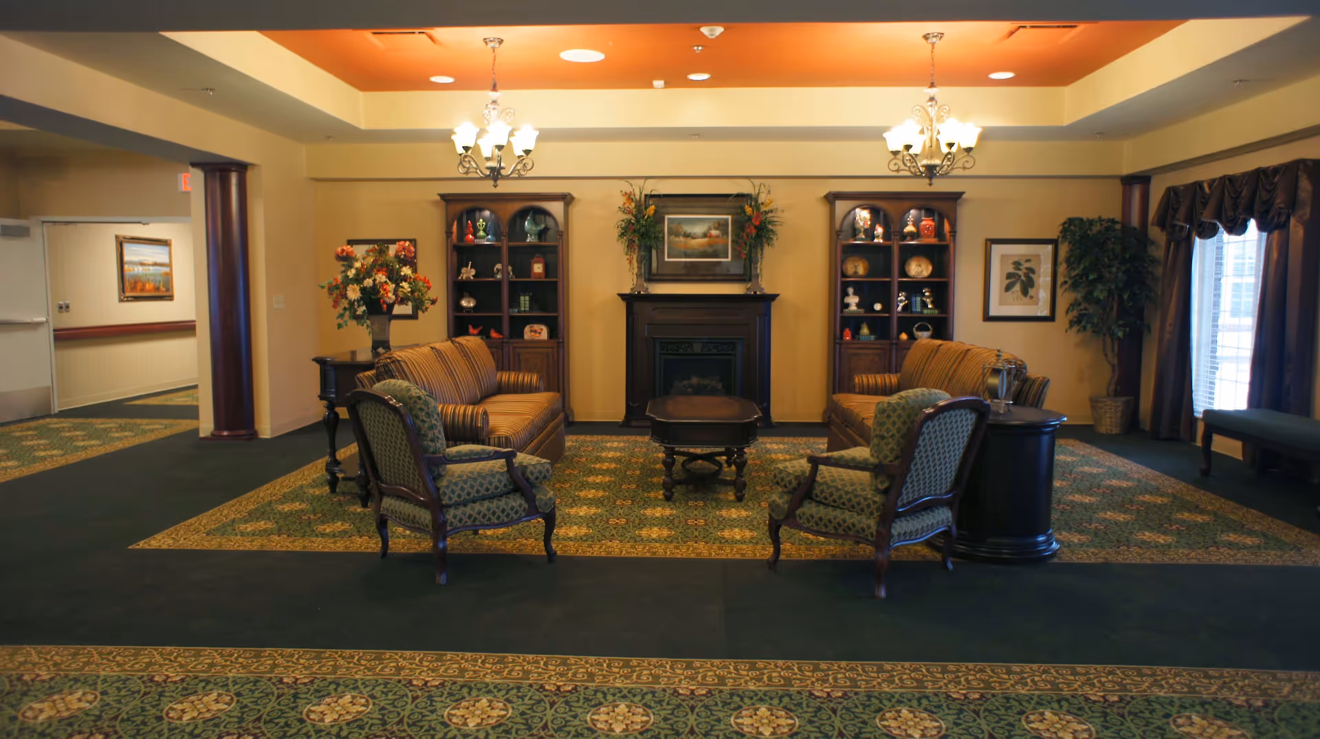A warmly lit common room with sofas and upholstered chairs arranged around a central coffee table and fireplace, flanked by bookcases and decorative plants.