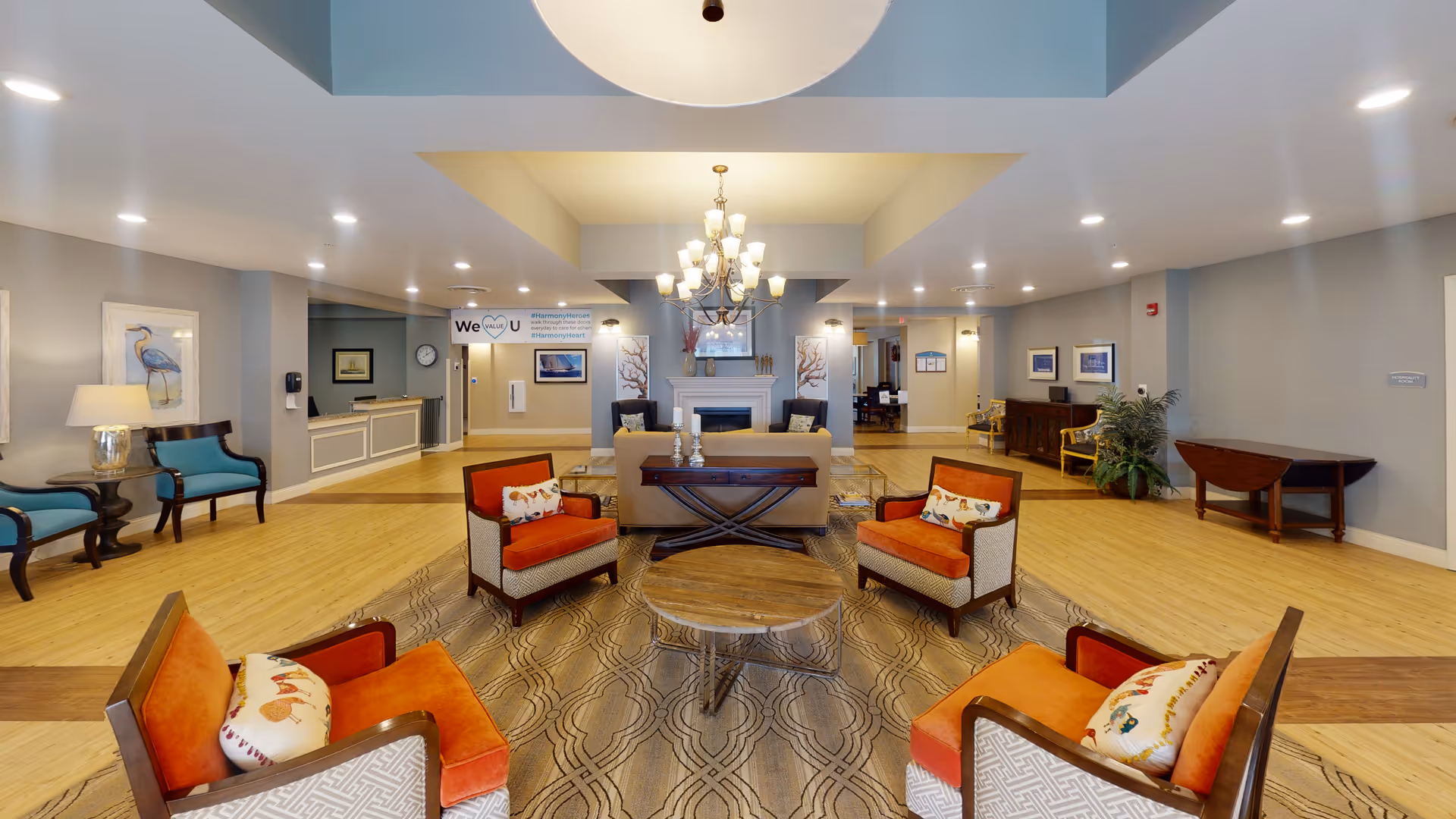 Spacious senior living facility lobby with seating arranged around a central coffee table, chandelier, and a reception desk in the background.