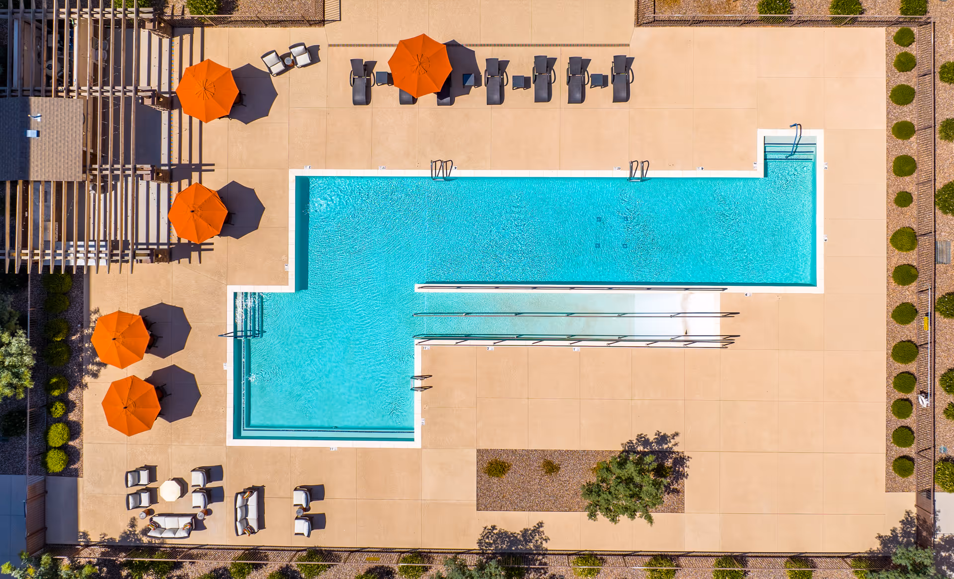 Aerial view of a rectangular outdoor swimming pool with a beige concrete deck surrounding it. The pool area features several orange umbrellas providing shade over lounge chairs and seating areas. There are also some plants and landscaping around the pool deck.