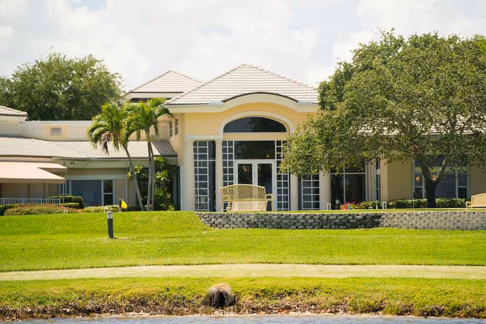 Exterior view of Sandhill Cove Retirement Living building with a well-maintained lawn, palm trees, and a large tree. The building features large windows and a light-colored roof with a bench on a raised stone platform in front.