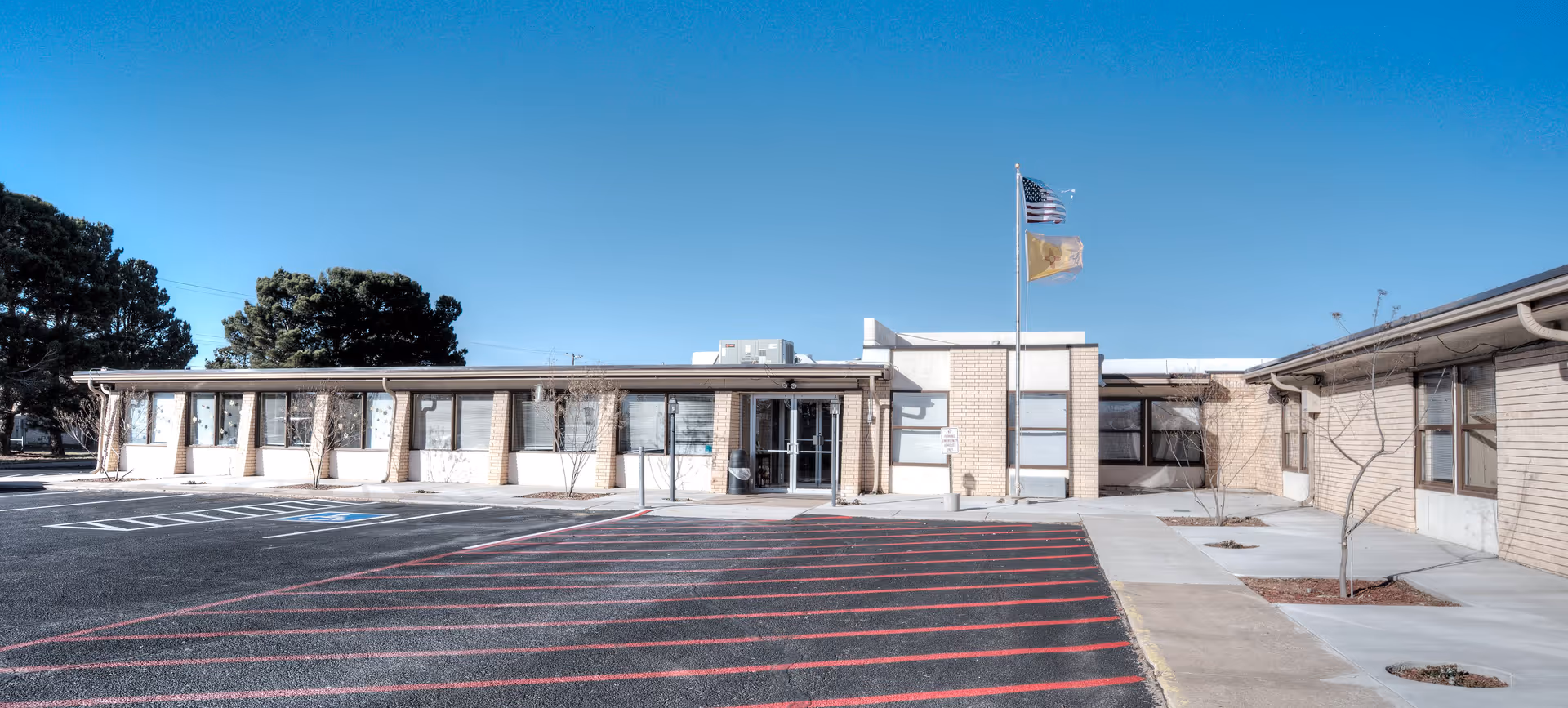 Exterior view of a single-story healthcare and rehabilitation center building with a parking lot in front. The building has light-colored brick walls, several windows, and two flagpoles displaying the American flag and another flag. The sky is clear and blue.