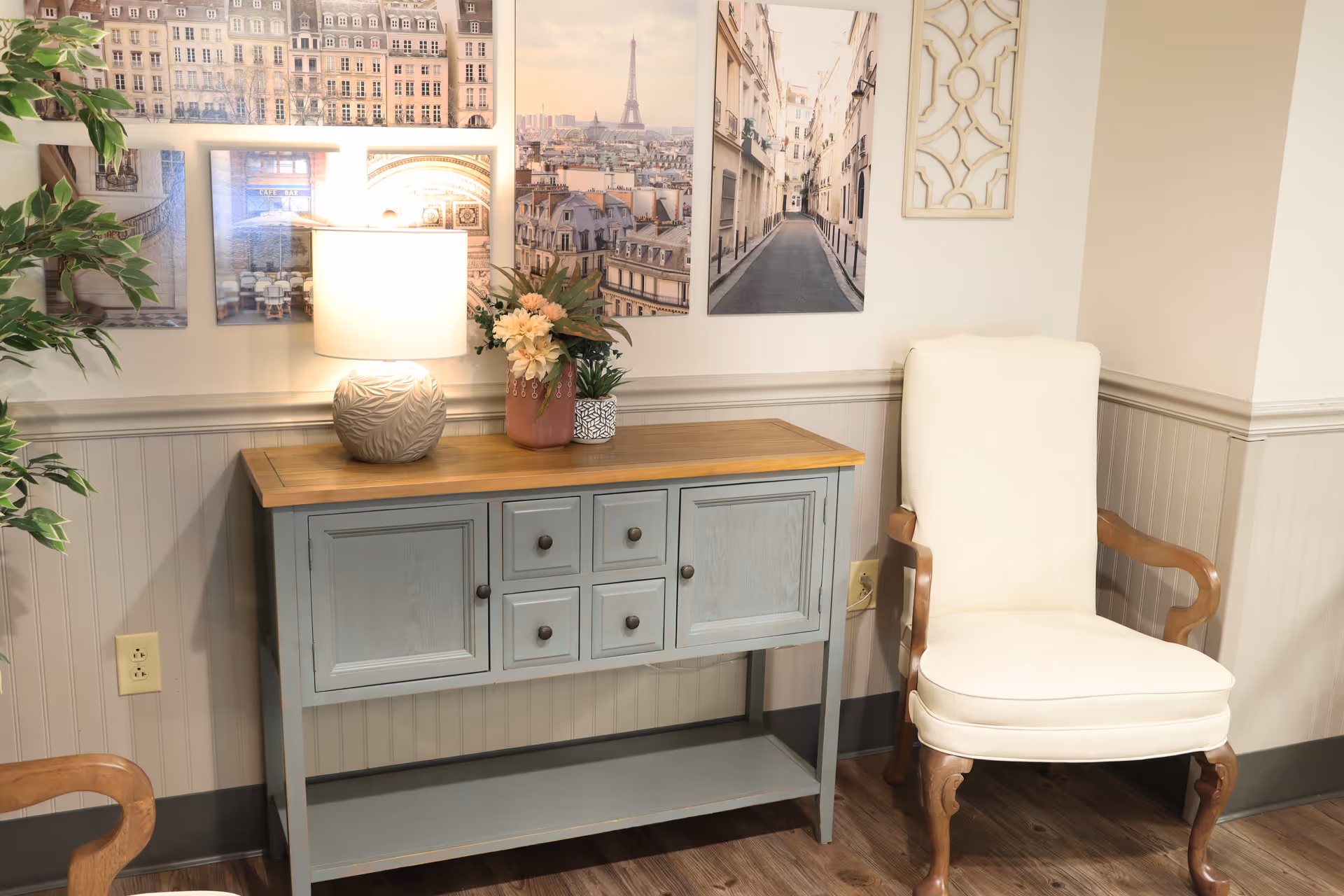 A cozy corner of a room featuring a light blue wooden sideboard with drawers and cabinets, topped with a decorative lamp and flower arrangements. Next to it is a cream-colored upholstered armchair with wooden armrests and legs. The wall behind has several framed pictures of Parisian scenes and a decorative wall hanging. The floor is wooden, and there is a green leafy plant partially visible on the left side.