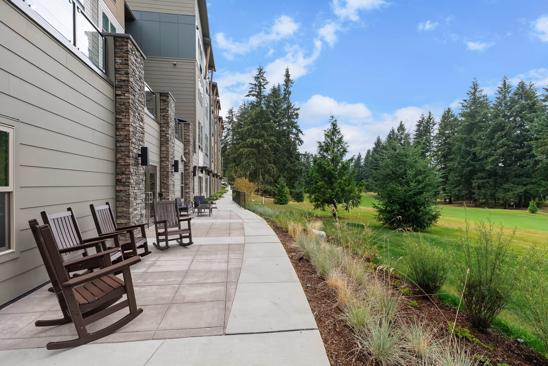 Sidewalk patio beside a senior living building with rocking chairs facing a landscaped lawn and trees.