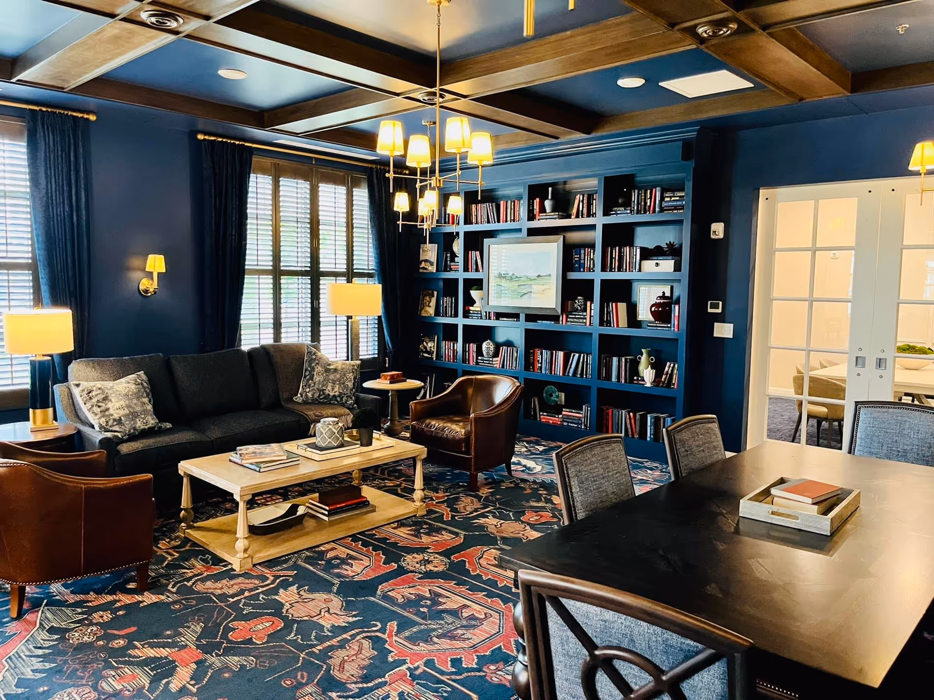 A cozy living room with dark blue walls and ceiling beams, featuring a large bookshelf filled with books and decorative items. The room has a dark gray sofa with patterned pillows, two brown leather armchairs, a wooden coffee table with books and decor, and a patterned rug. There is a dining table with chairs in the foreground and a set of glass double doors leading to another room.