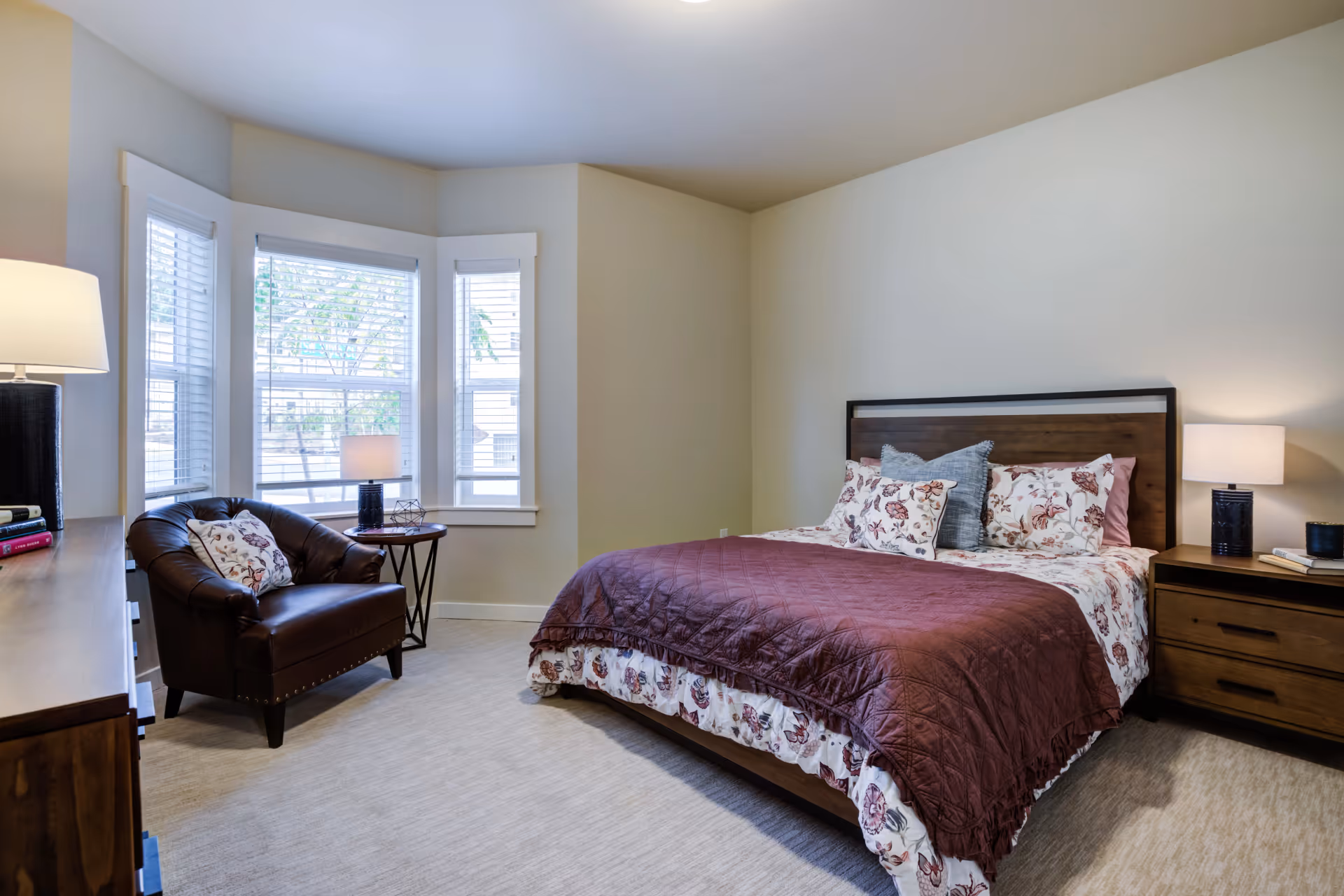 A cozy bedroom with a large bed featuring floral bedding and a maroon quilt. There is a wooden nightstand with a lamp on the right side of the bed. A dark brown leather armchair with a floral pillow sits near a bay window with white blinds. A wooden dresser with a lamp and books is on the left side of the room. The walls are painted light beige and the floor is carpeted.