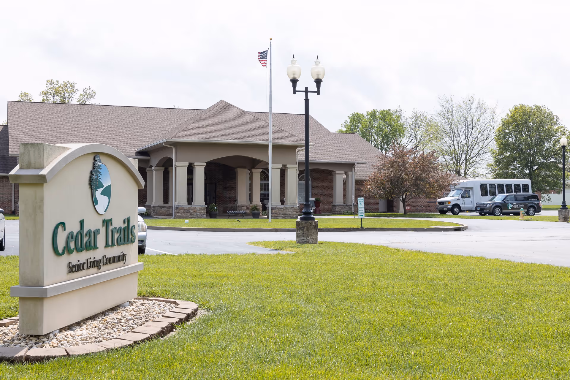 Exterior view of Cedar Trails Senior Living Community building with a large sign in the foreground, a flagpole with an American flag, a street lamp, parked vehicles including a van and a car, and green grass and trees surrounding the area.