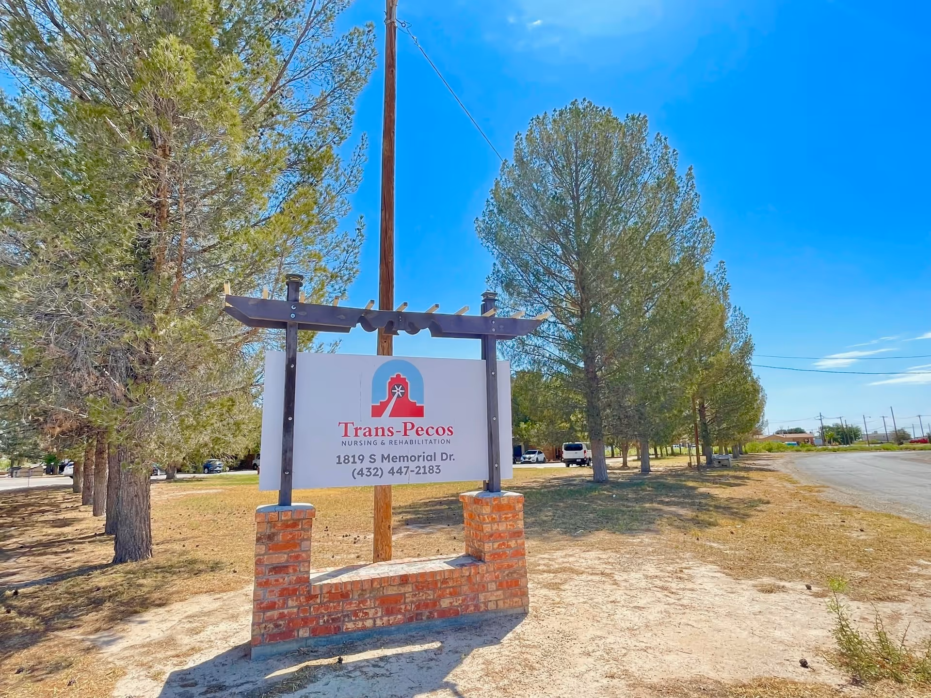 Outdoor view of a sign for Trans-Pecos Nursing & Rehabilitation located on a brick base with trees and a clear blue sky in the background along a roadside.