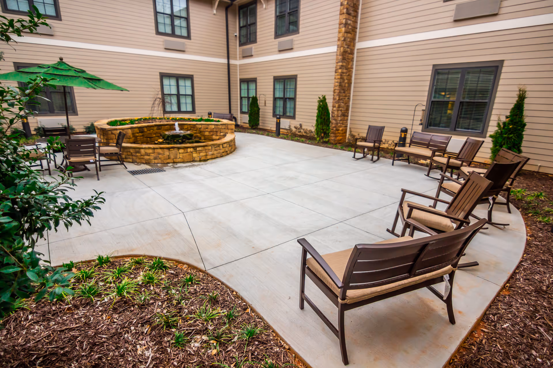 Outdoor courtyard area at Brickmont at Woodstock featuring a circular stone water fountain in the center, surrounded by multiple brown cushioned benches and chairs. The courtyard is paved with concrete and bordered by landscaped garden beds with small plants and mulch. The building exterior has beige siding with several windows and stone accents.