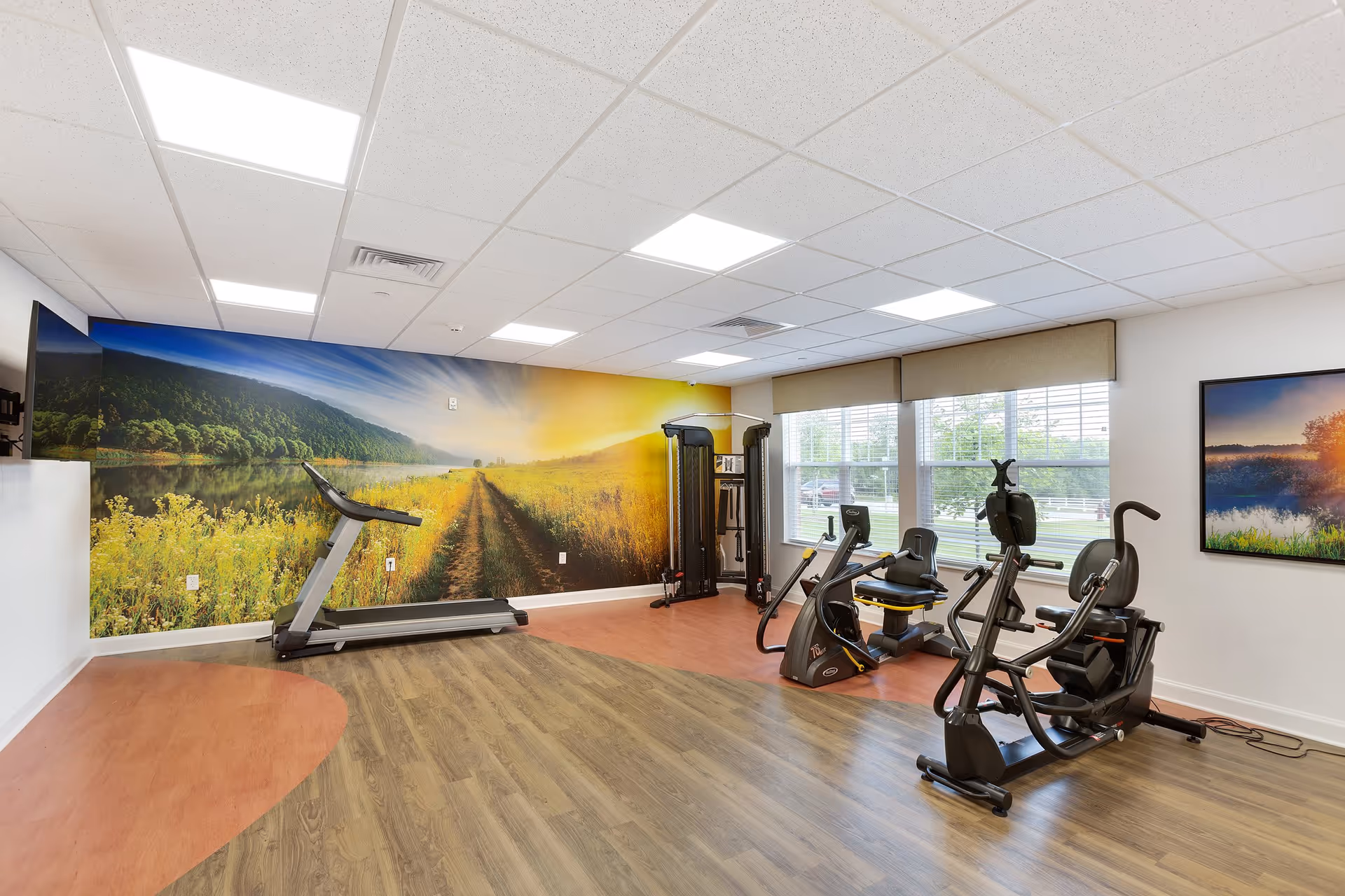 A bright fitness room with exercise equipment including a treadmill, a multi-function weight machine, and two recumbent exercise bikes. The room has wood and orange flooring, a large mural of a scenic outdoor path with yellow flowers on one wall, and large windows with beige blinds letting in natural light. A flat-screen TV is mounted on the wall.