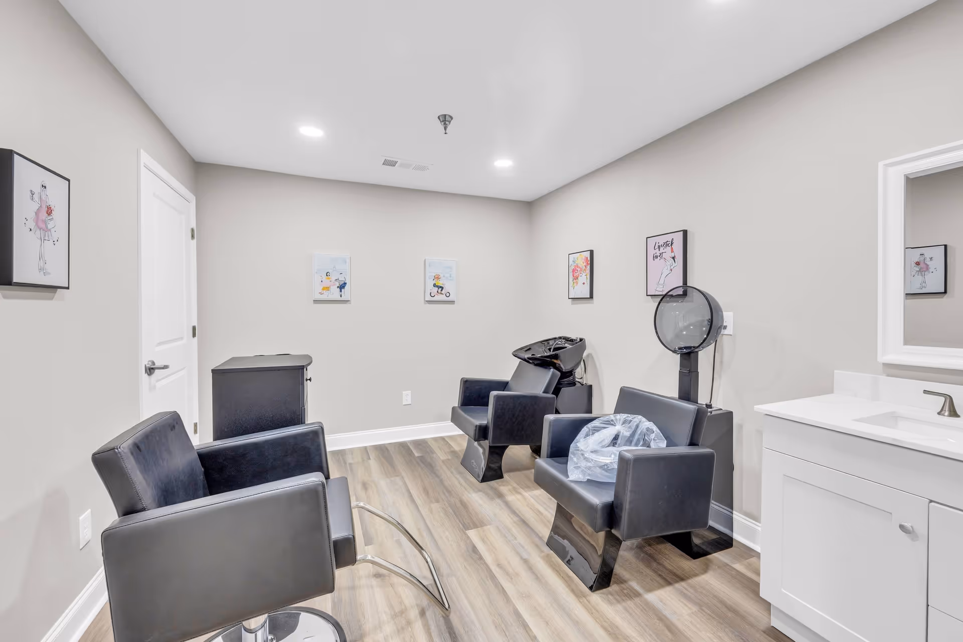 Interior view of a small salon area in a senior living facility with three black salon chairs, one with a plastic cover, a white sink with a mirror, light wood flooring, and framed artwork on beige walls.