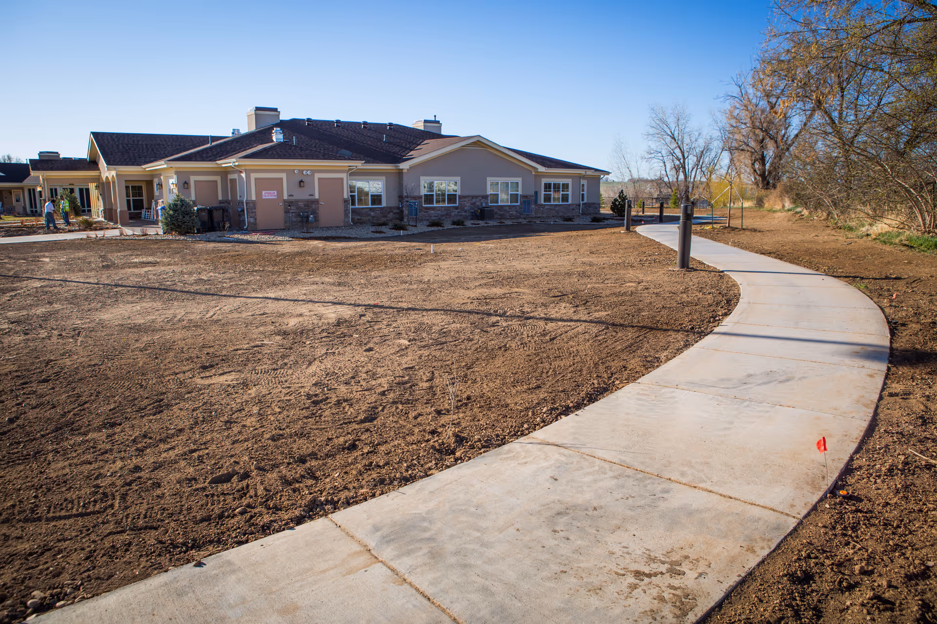 Curved concrete walkway leading to a single-story senior living building with bare landscaping under a clear blue sky.