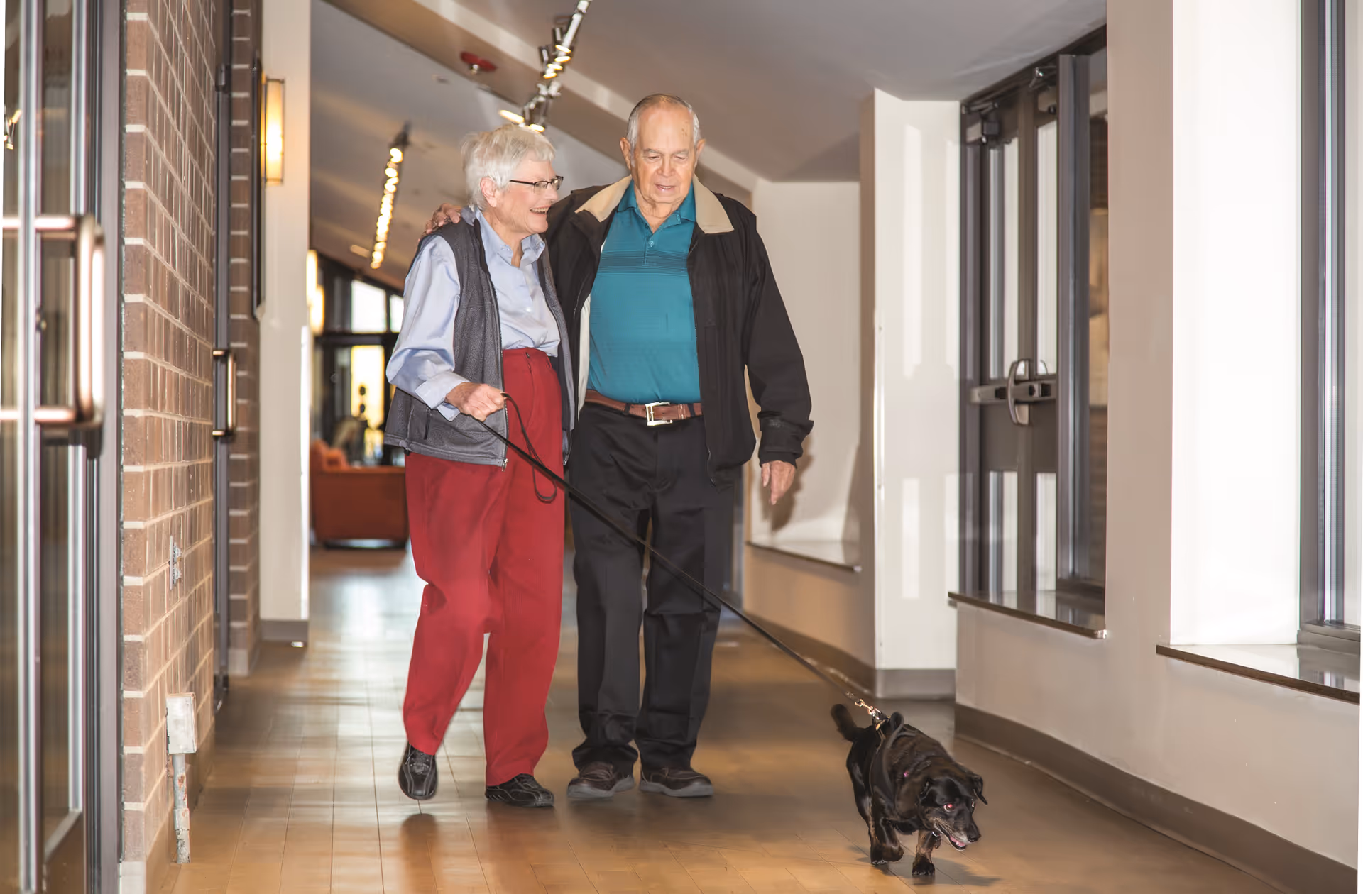 An elderly couple walking down a hallway inside a senior living facility, with the woman holding a leash and walking a small black dog. The man has his arm around the woman, and they are both smiling. The hallway has wooden flooring, brick and white walls, and large windows on one side.