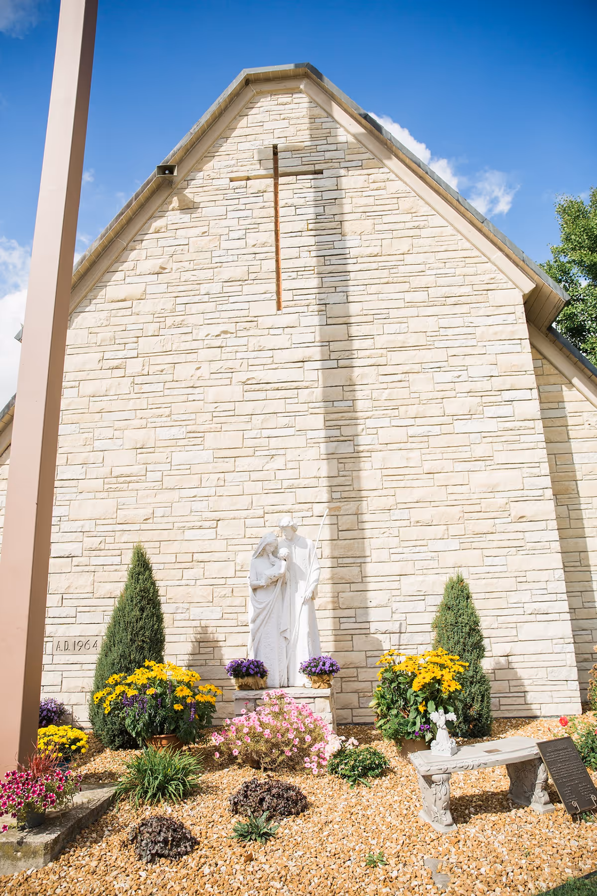 Outdoor garden area with a stone wall building in the background. A white statue of a religious family figure is centered, surrounded by colorful flowers and small bushes. There is a stone bench and a plaque on the right side. The sky is blue with some clouds.