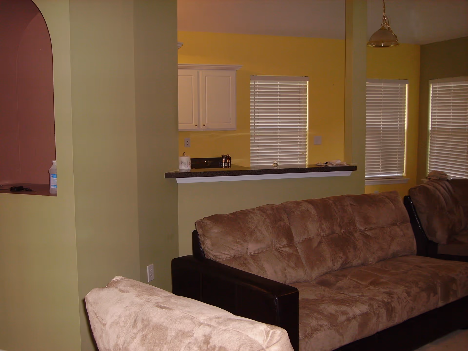 Interior view of a living room area with beige cushioned sofas, yellow walls, and windows with white blinds. A partial view of a kitchen with white cabinets and a countertop is visible in the background.