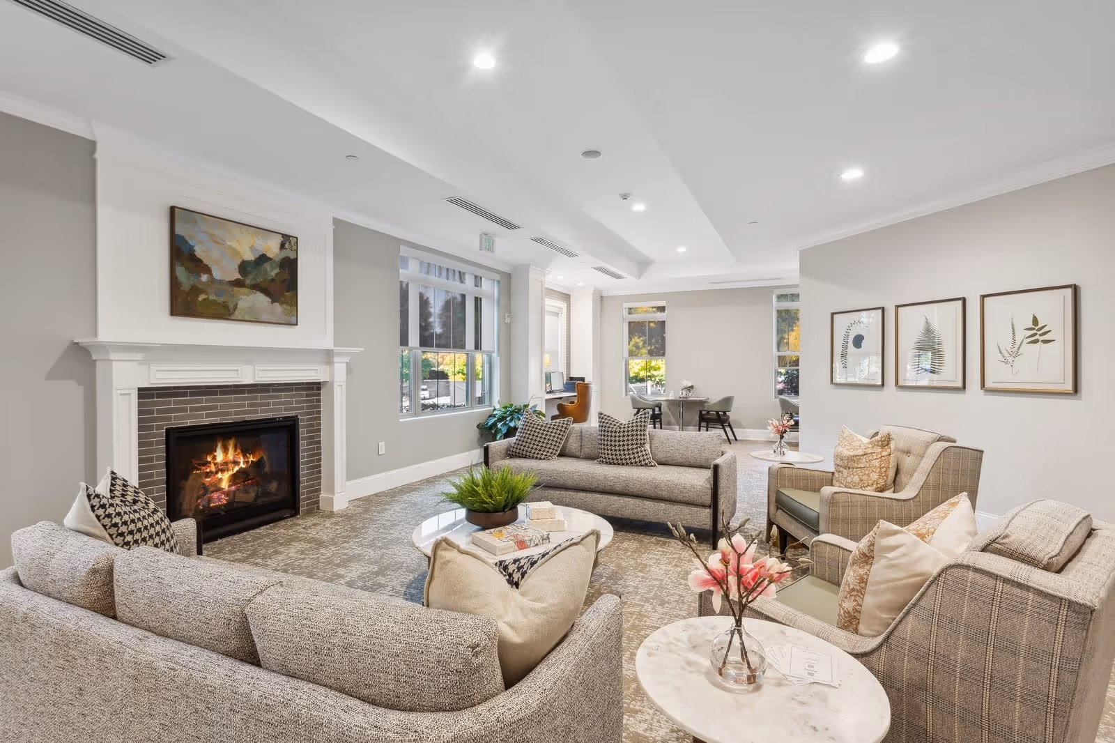 A bright and cozy senior living facility common area with a lit fireplace, two gray sofas, two armchairs, a round marble coffee table with a vase of pink flowers, and framed botanical artwork on the wall. Large windows let in natural light, and a small table with chairs is visible in the background.