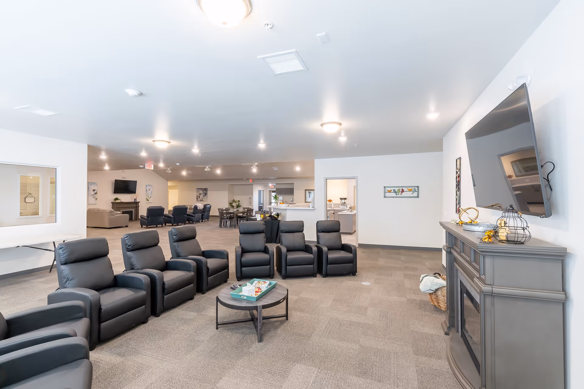 A spacious senior living facility common area with a row of black recliner chairs arranged in a semi-circle facing a wall-mounted flat screen TV above a gray electric fireplace. In the background, there are additional seating areas with sofas and chairs, a dining table, and a kitchen area. The room has white walls, carpeted floors, and bright ceiling lights.