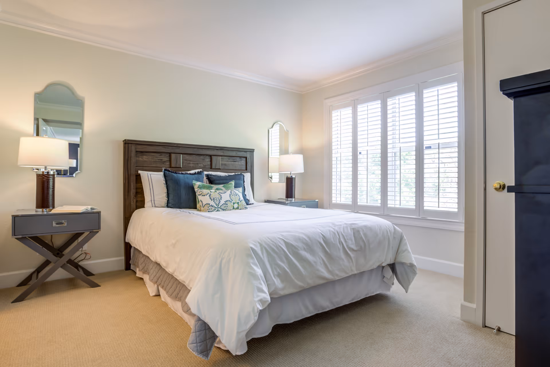 A bright and neatly arranged bedroom with a large bed featuring white bedding and decorative pillows. The room has beige walls, carpeted floor, two bedside tables each with a lamp and a mirror above it, and a large window with white plantation shutters allowing natural light to enter.