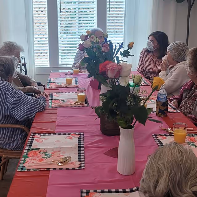 A group of elderly people and a caregiver wearing a mask sitting around a dining table covered with a pink tablecloth. The table is set with floral placemats, glasses of orange juice, and vases with colorful roses. Sunlight streams through white blinds in the background.