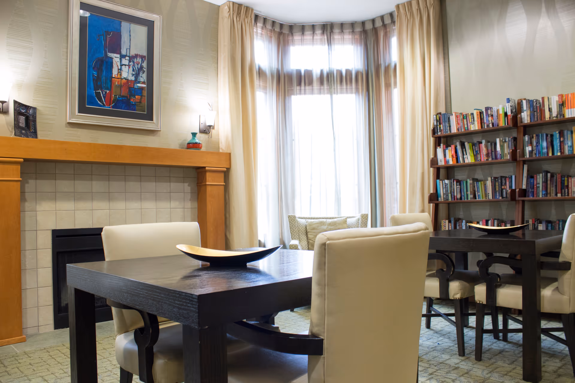 A cozy interior room with two dark wooden tables surrounded by cream-colored upholstered chairs. There is a fireplace with a wooden mantle and a colorful abstract painting above it. A large window with sheer curtains allows natural light to fill the room. A bookshelf filled with books is visible on the right side of the room.