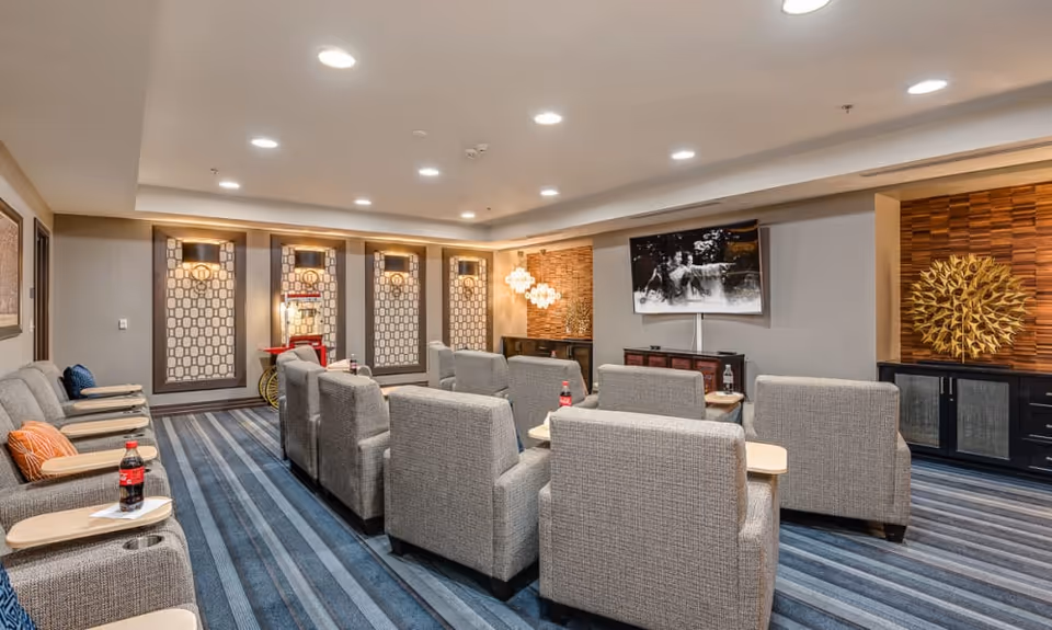 A cozy senior living common area with multiple gray upholstered armchairs arranged in rows facing a wall-mounted flat-screen TV. The room features blue striped carpeting, decorative wall panels with lighting, a popcorn machine in the corner, and modern wall art including a golden sunburst sculpture. The ceiling has recessed lighting providing bright illumination.