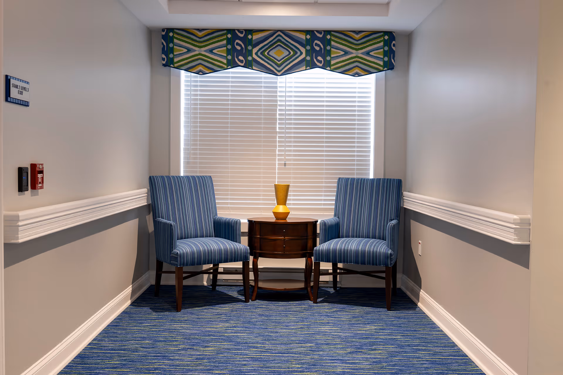 A small seating area with two blue striped armchairs positioned on either side of a round wooden side table with a yellow vase on top, set against a window with white blinds and a colorful valance. The area has blue patterned carpet and light gray walls with white trim and a handrail.