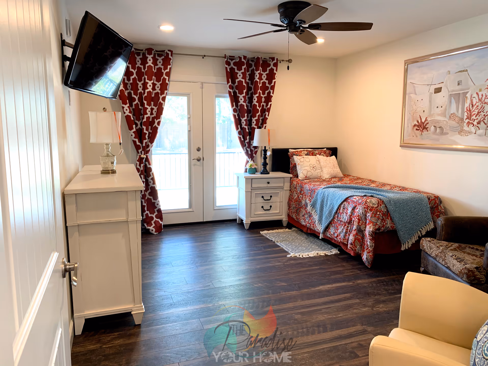 A cozy bedroom with a single bed covered in a red floral bedspread and a blue throw blanket. There is a white nightstand with a lamp and a small flower arrangement next to the bed. A white dresser with a lamp is positioned against the left wall, and a flat-screen TV is mounted above it. The room has dark wooden floors, red patterned curtains on glass double doors, a ceiling fan with lights, and a framed painting on the wall. There is also a brown armchair and a yellow chair partially visible in the foreground.