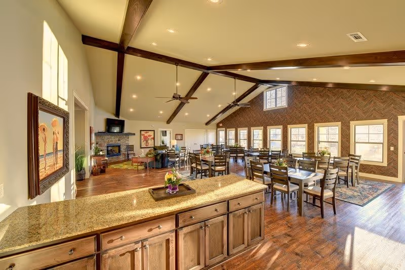 Spacious and well-lit common area with wooden floors and exposed ceiling beams. The room features multiple dining tables with chairs, a cozy seating area with a fireplace and TV, and large windows letting in natural light. A granite countertop with wooden cabinets is visible in the foreground.