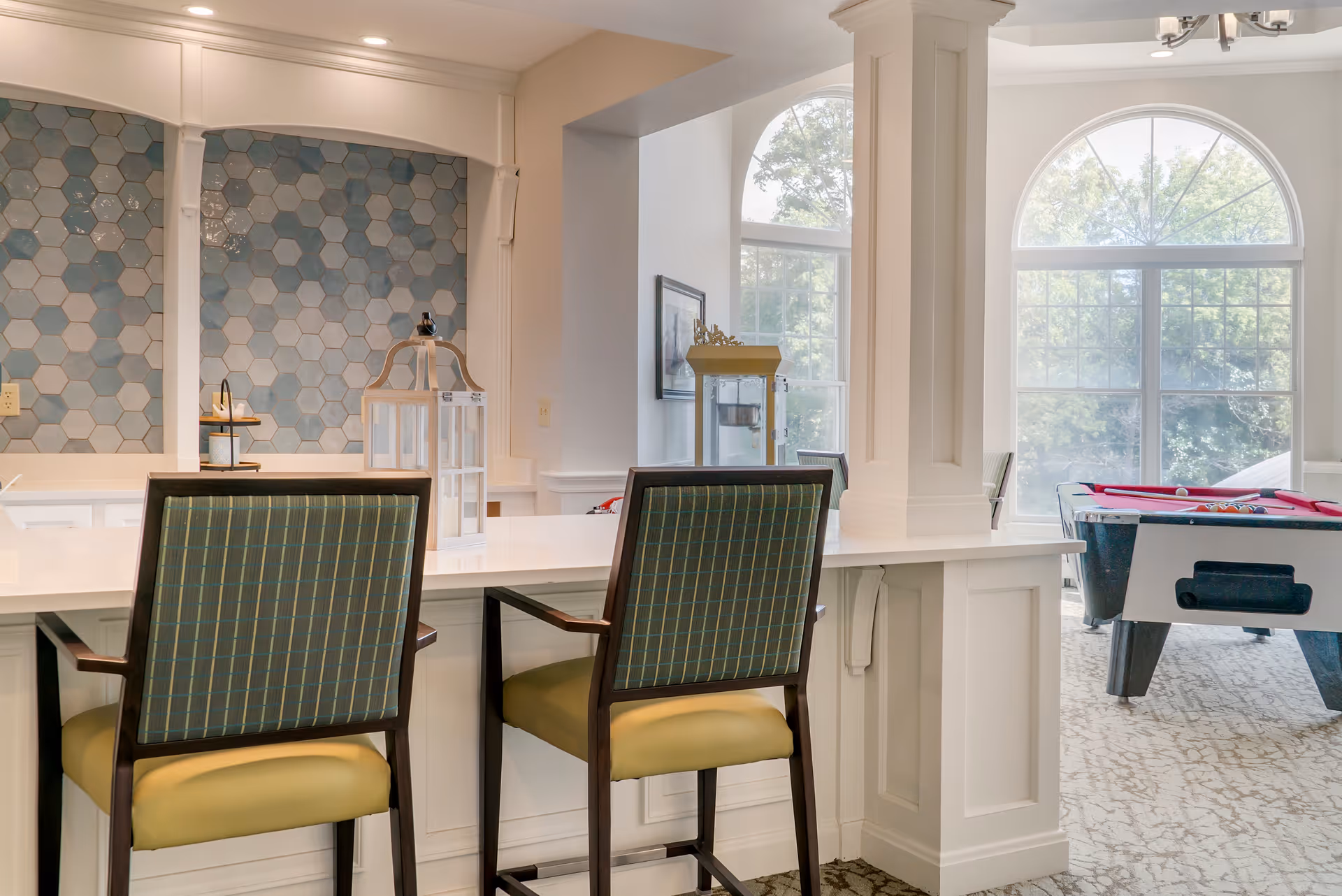 Interior view of a senior living facility common area with two chairs at a white counter. The background features a wall with hexagonal blue and gray tiles, large arched windows letting in natural light, and a pool table with a red cover.