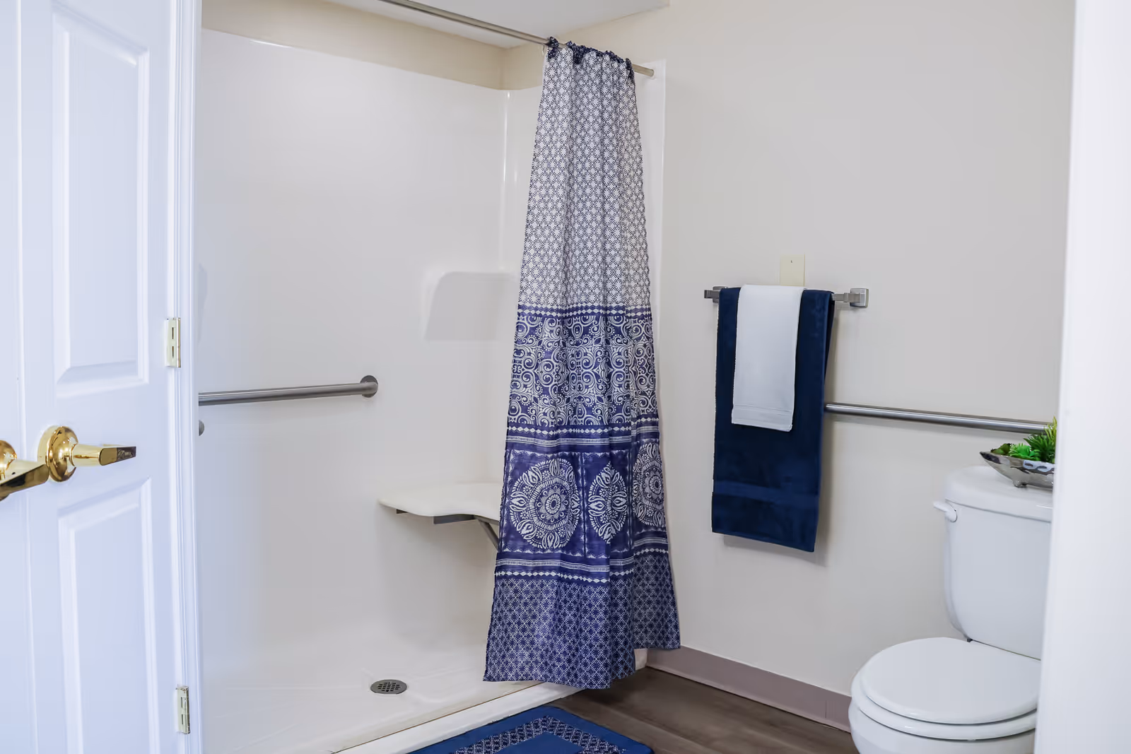 Accessible bathroom with a walk-in shower and built-in seat, blue patterned shower curtain, grab bars, toilet and towel on a rack.
