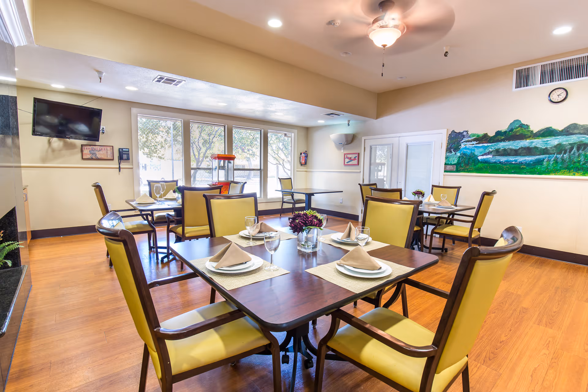 A bright dining room with wooden floors and multiple tables set with plates, glasses, and folded napkins. The chairs have yellow cushions and dark wooden frames. Large windows let in natural light, and a ceiling fan with a light fixture is on the ceiling. A colorful landscape painting is on one wall, and a flat-screen TV is mounted on another wall.