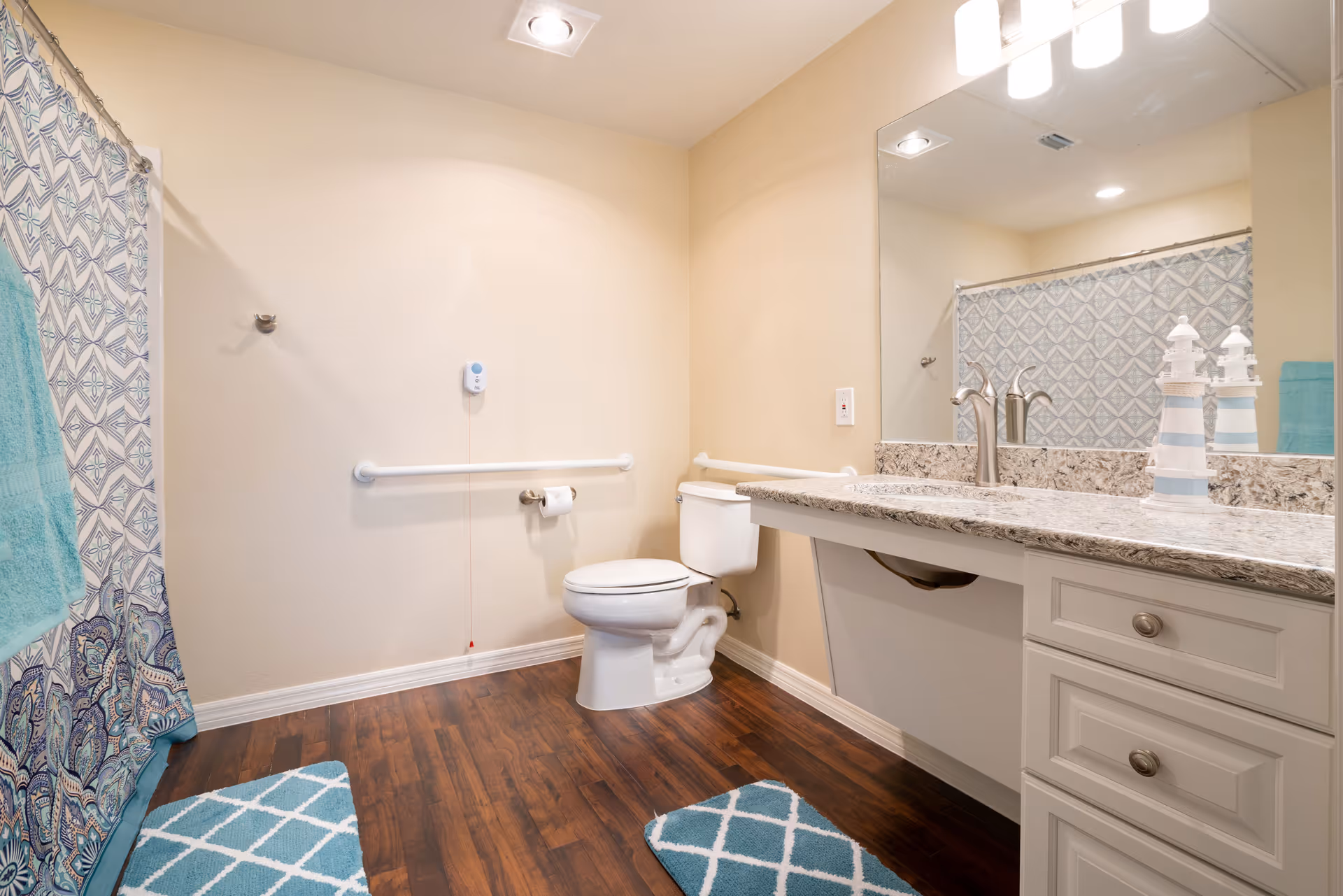 A clean and spacious bathroom with a toilet, a large mirror above a granite countertop with a sink, and a shower with a patterned curtain. The floor is wooden with two blue and white patterned rugs. There are grab bars near the toilet and a pull cord for assistance. The walls are painted beige.