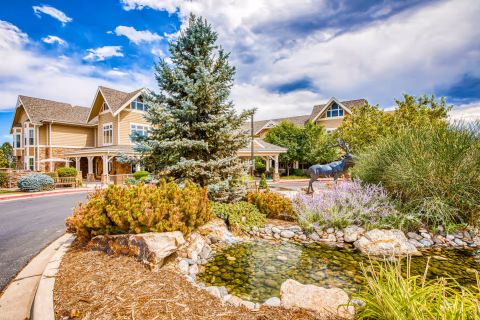 Exterior view of MorningStar Assisted Living & Memory Care at Applewood showing a landscaped garden with a small pond, rocks, shrubs, a large evergreen tree, and a metal sculpture of a deer. The building with beige siding and multiple windows is visible in the background under a partly cloudy sky.