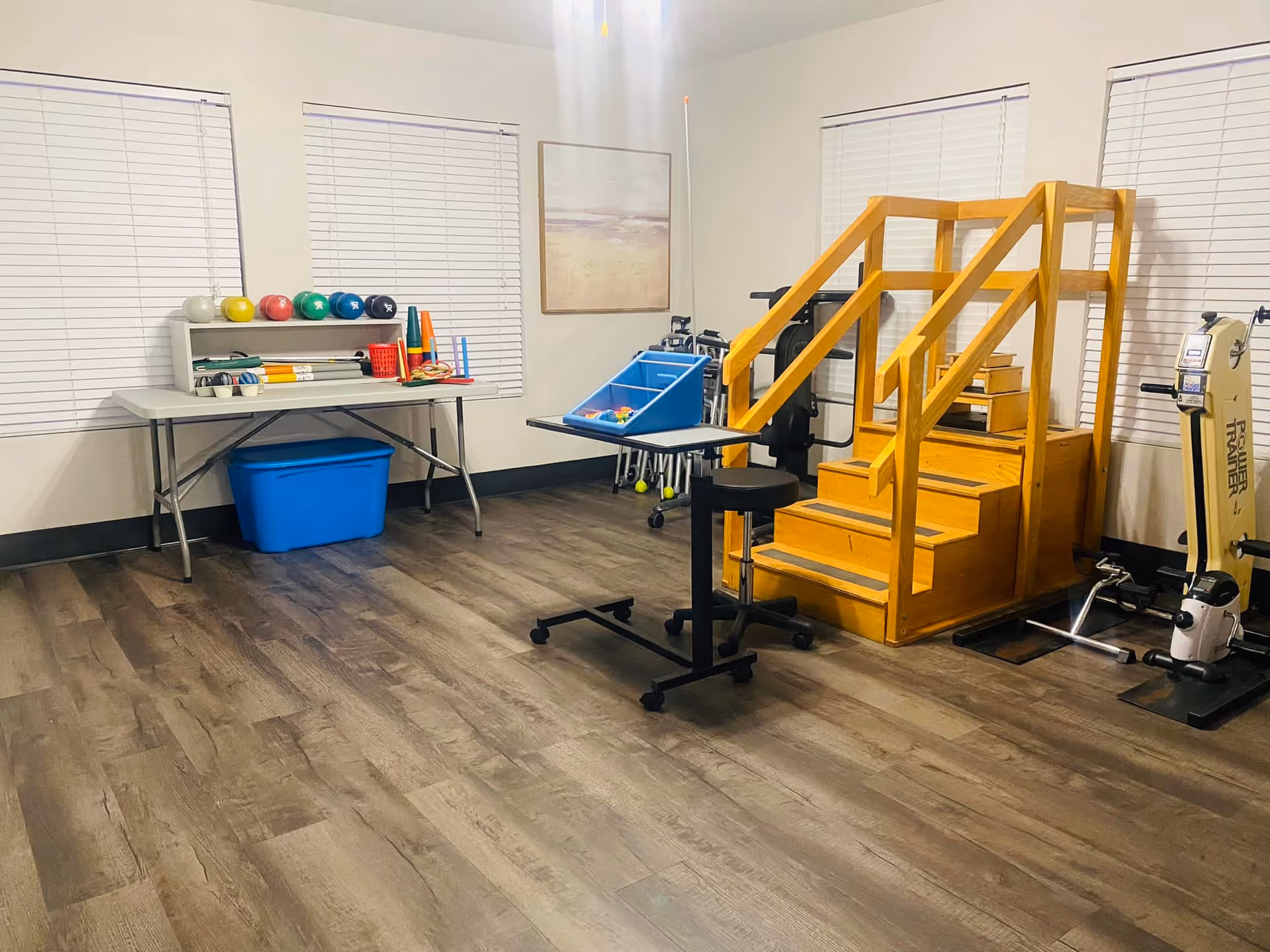 A rehabilitation therapy room with wooden stairs for physical therapy, exercise equipment including a rowing machine, a table with colorful therapy balls and cones, and a blue storage bin underneath the table. The room has three windows with white blinds and a framed picture on the wall.