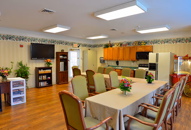 A bright and spacious common area in a senior living facility featuring a long table covered with a beige tablecloth surrounded by green cushioned chairs. The room has wooden flooring, a wall-mounted flat screen TV, a small bookshelf, and a kitchenette with wooden cabinets, a refrigerator, microwave, and stove. There are floral arrangements on the table and a popcorn machine in the corner. The walls have a floral wallpaper border near the ceiling.