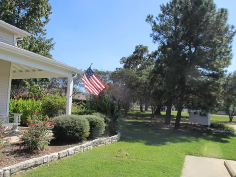 View of a green lawn and garden area with bushes and flowers next to a building porch displaying an American flag. Trees and a small white shed are visible in the background under a clear blue sky.