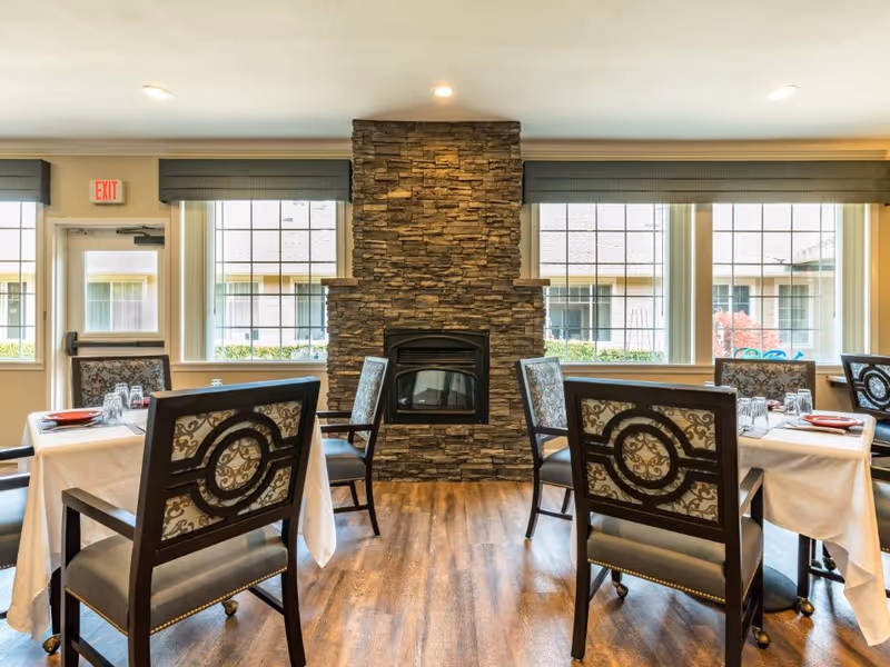 Dining room with set tables and upholstered chairs centered around a stone fireplace beneath large windows.