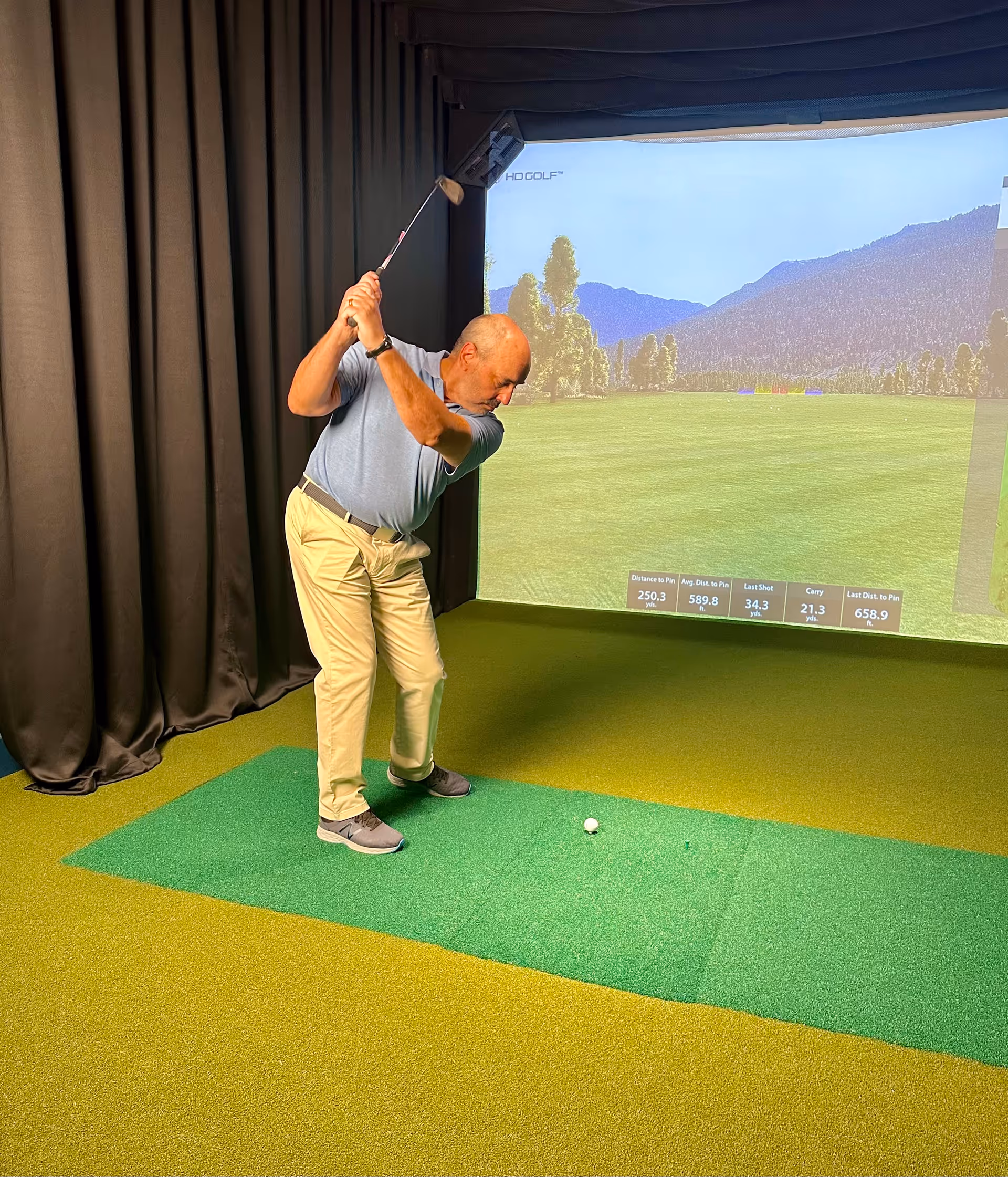 An elderly man is playing indoor golf using a golf simulator. He is mid-swing, standing on a green mat with a golf ball on a tee in front of him. The simulator screen shows a golf course with trees and mountains in the background.